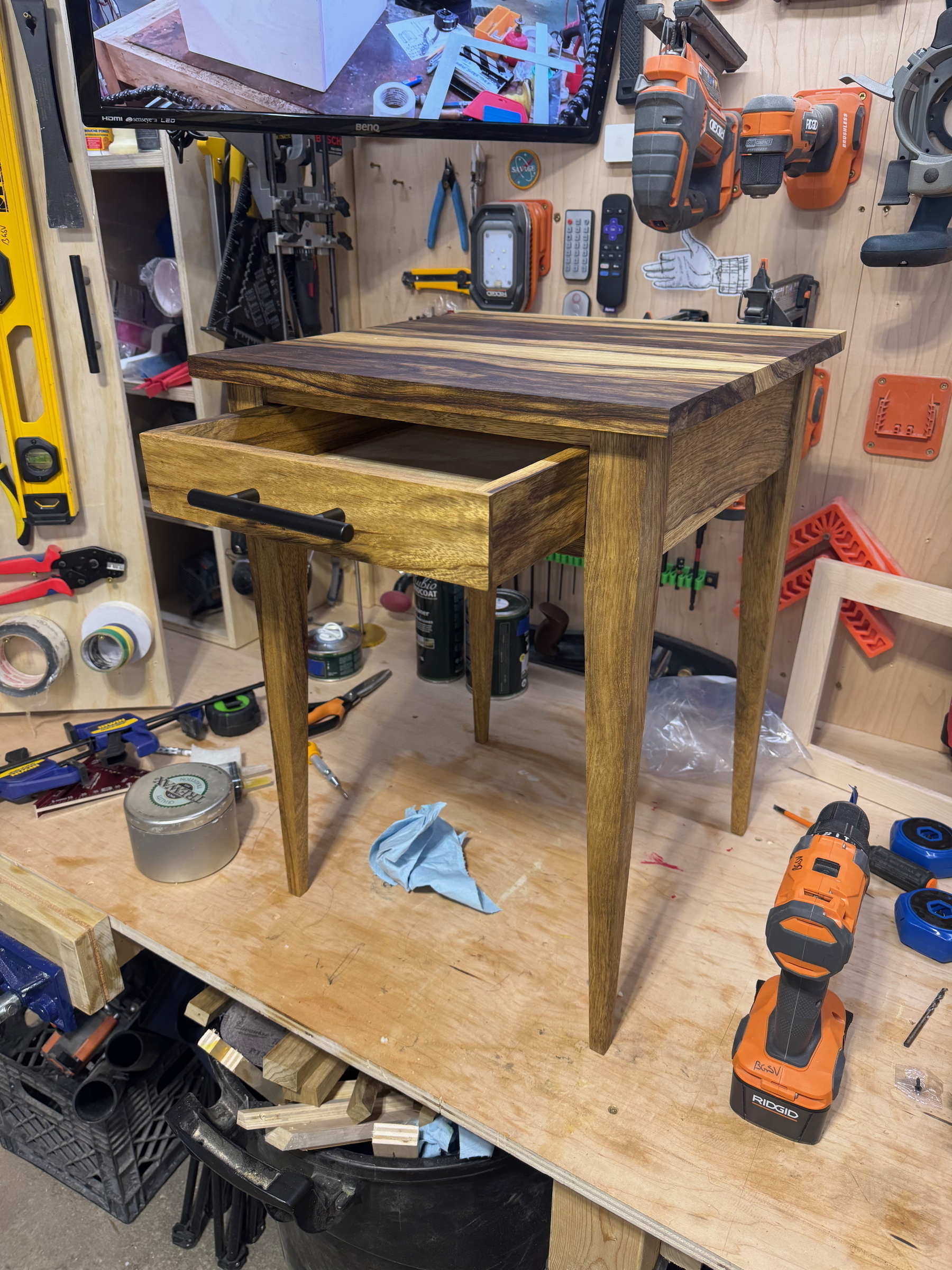 A wooden table with a drawer is displayed in a workshop surrounded by various tools.