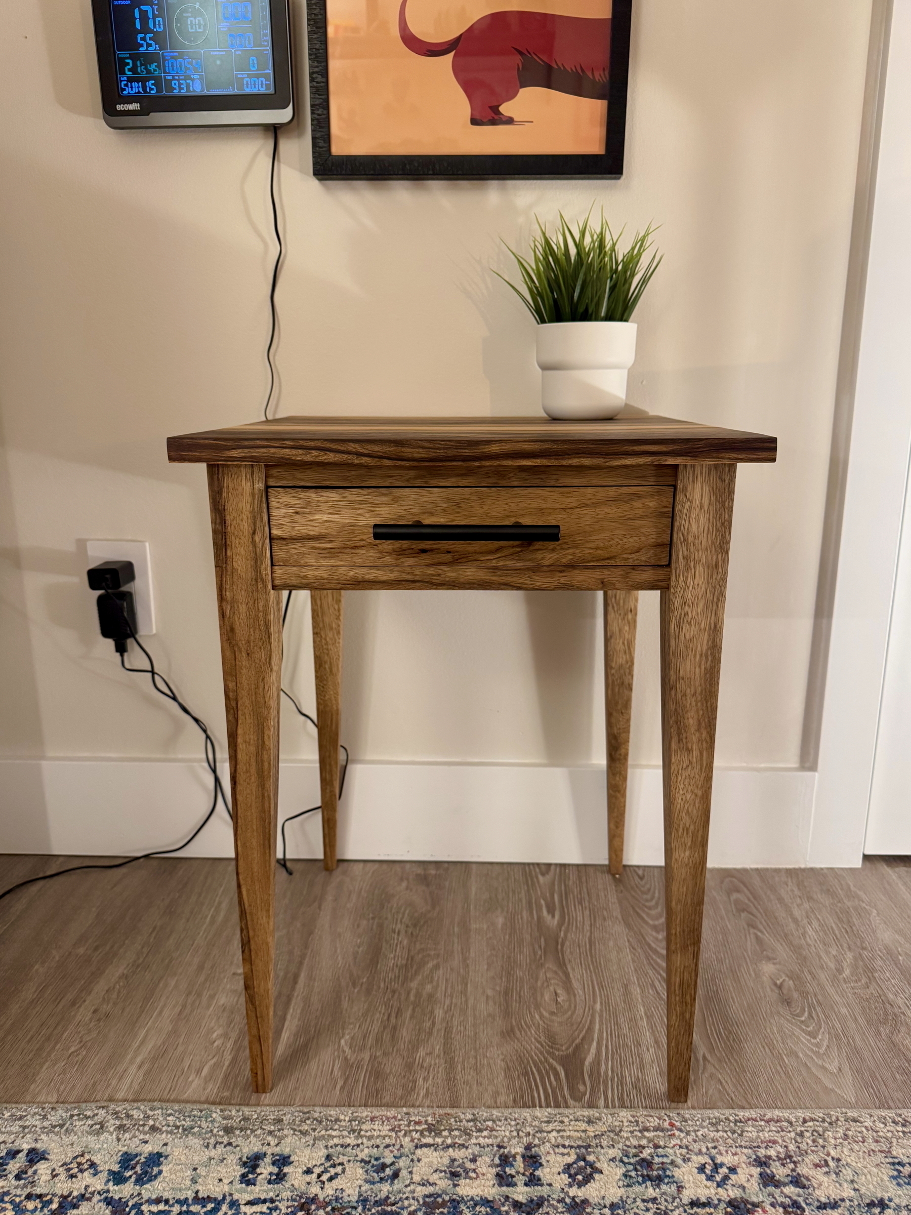 A wooden side table with a drawer, topped with a small potted plant, stands against a wall adorned with a colorful framed picture and a digital clock.