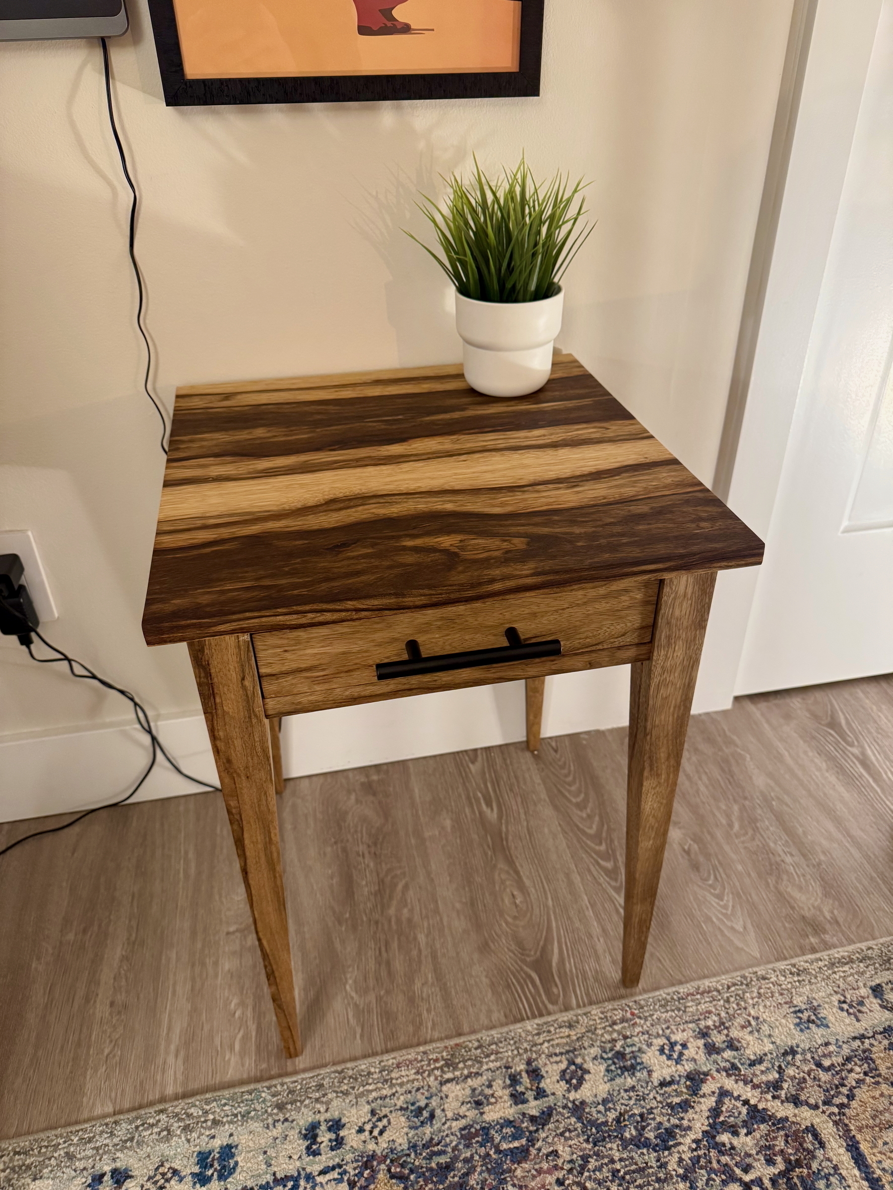 A wooden side table with a drawer and a potted plant on top is placed on a hardwood floor.